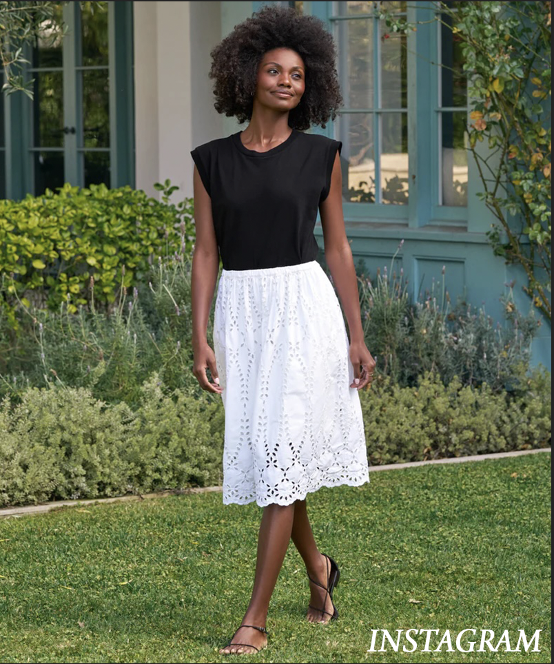 A woman with curly hair walks on grass, wearing a black sleeveless top, a white knee-length eyelet skirt, and sandals. She is outside near a building with large windows and green plants. The word "INSTAGRAM" is in the corner. Fearrington Village